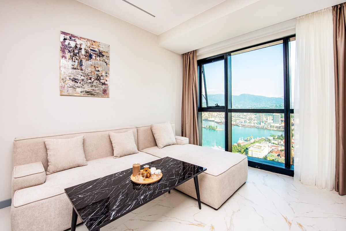 A modern living area features a light-colored sectional sofa complemented by soft cushions. A sleek black coffee table is arranged with snacks, and a piece of abstract artwork adorns the wall. Floor-to-ceiling windows showcase panoramic views of the surrounding landscape.