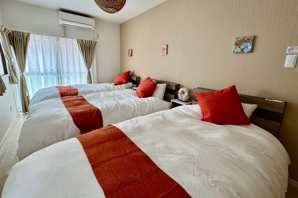 The bedroom features three spacious beds dressed in crisp white linens, each adorned with vibrant orange accent pillows. A large window with sheer curtains allows natural light, while subtle artwork adds character to the beige walls.
