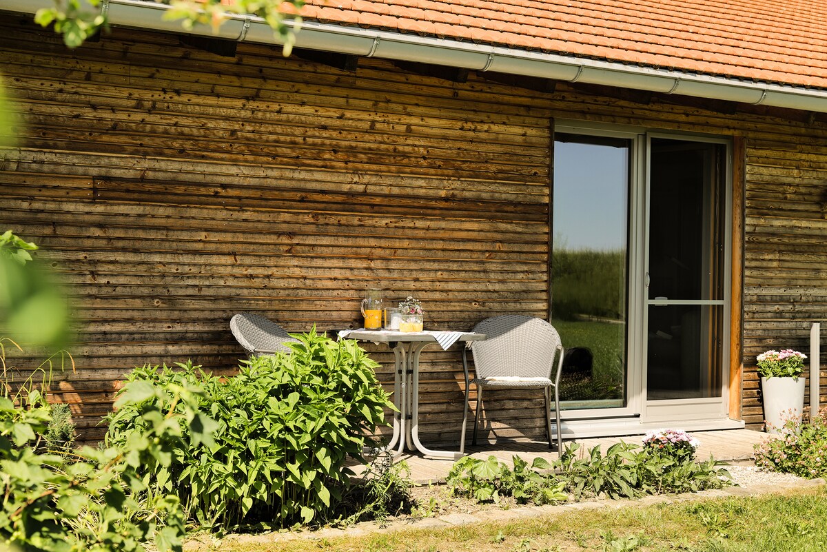 A cozy outdoor terrace is presented, featuring a small round table with two chairs. Refreshing drinks can be seen atop the table. The background includes a well-maintained wooden wall and vibrant green plants, enhancing the peaceful ambiance of the space.