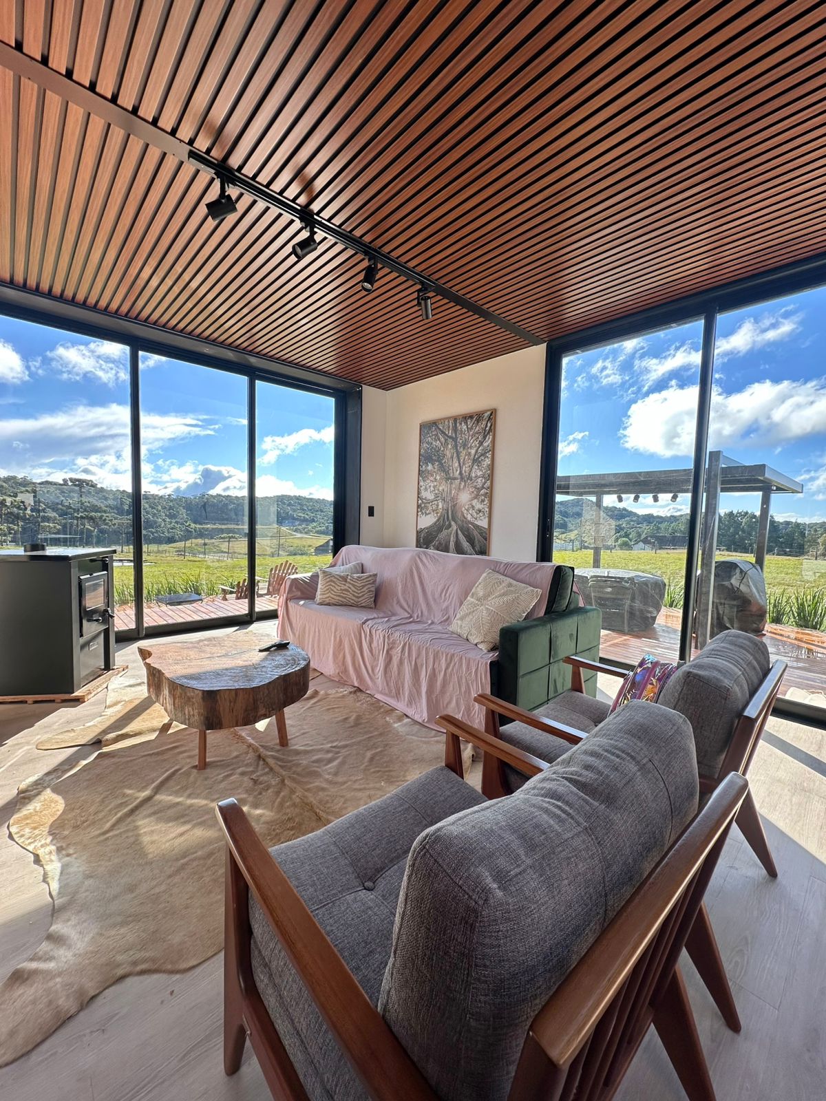 A sunlit living area features large glass panels that provide a panoramic view of the surrounding nature. A comfortable sofa, two armchairs, and a wooden coffee table are arranged on a soft rug. A piece of artwork is displayed on the wall, enhancing the space.