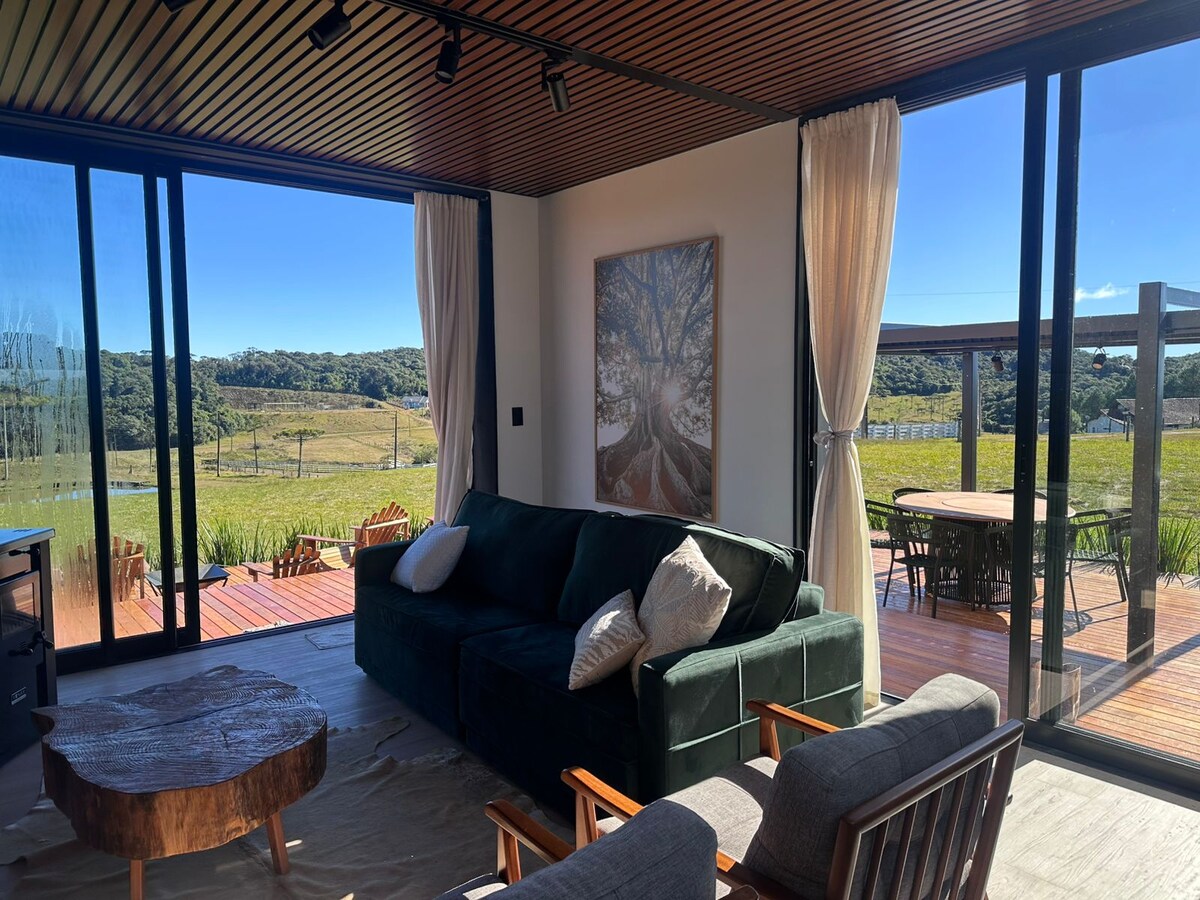 A modern living area is showcased, featuring large glass doors that open to a scenic view of nature. A comfortable green sofa is placed beside a wooden coffee table, while two armchairs provide additional seating. Natural light illuminates the space, accentuated by soft curtains.