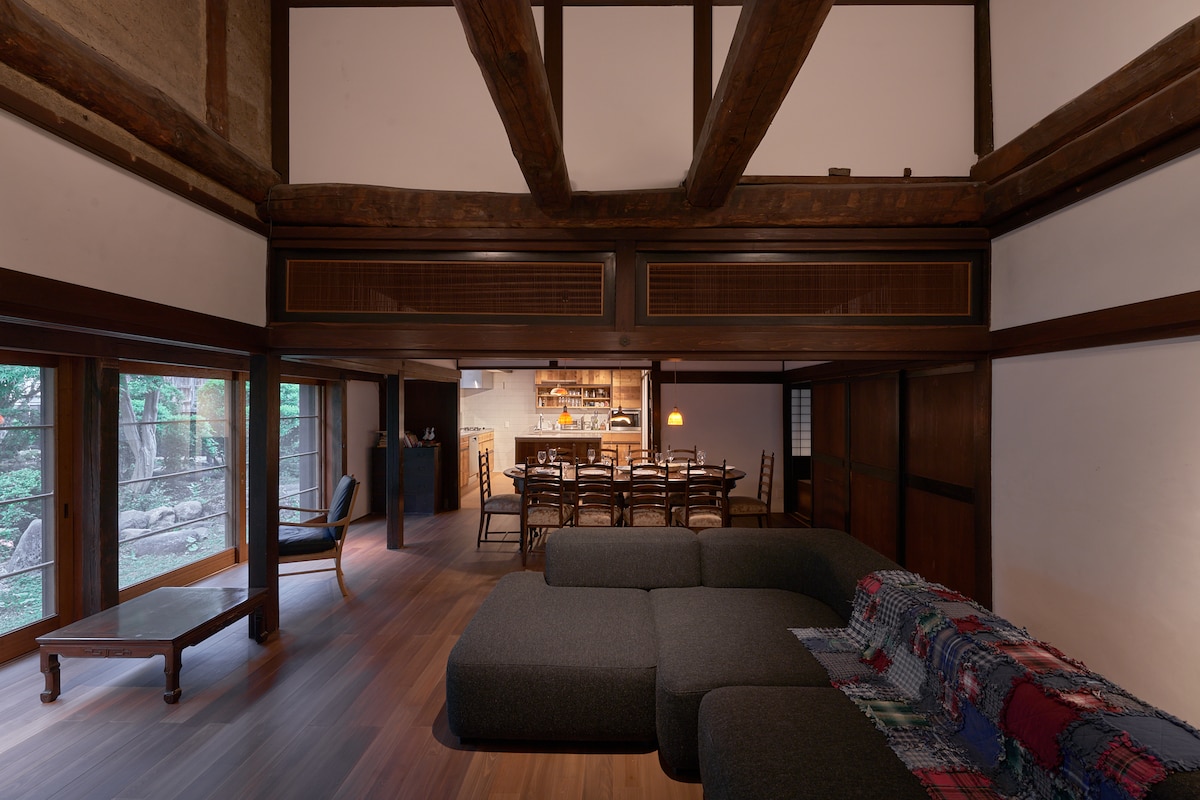 An open living area showcases a spacious, modern sofa positioned beside a wooden coffee table. High wooden beams complement the traditional architecture, while a dining table can be seen in the background, illuminated by soft lighting. Large windows offer views of the surrounding greenery.