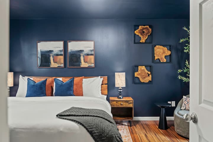 Elegante dormitorio de invitados con paredes de color azul marino intenso, arte abstracto moderno y ricos detalles en madera.  Mesitas de noche de madera a juego e iluminación cálida, decoración de pared de madera orgánica y un acogedor rincón de lectura completan el espacio sofisticado y acogedor. Cama tamaño king.