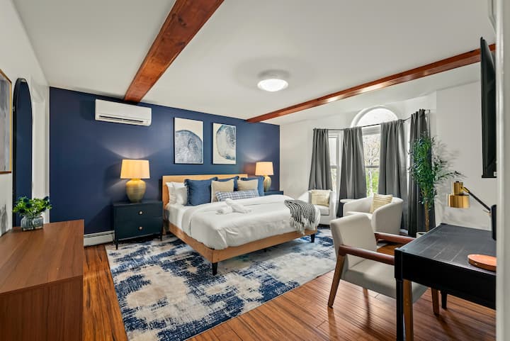 El amplio dormitorio principal cuenta con una llamativa pared decorativa azul marino, vigas de madera a la vista en el techo y una cama tamaño king con texturas acogedoras. La luz natural entra a través de una gran ventana arqueada, complementando el moderno espacio de trabajo y la acogedora sala de estar.