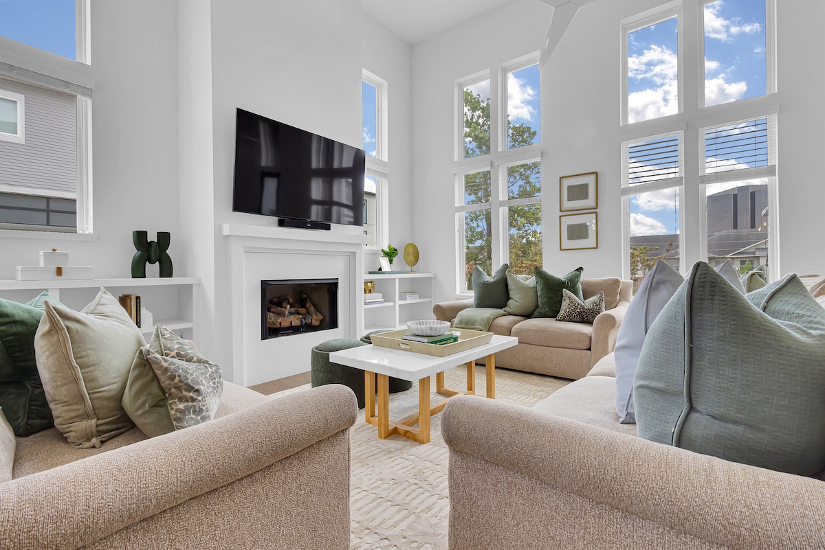 The spacious living area features large windows allowing natural light to fill the room. Comfortable sofas adorned with various throw pillows are positioned around a modern coffee table. A mounted flat-screen TV and a decorative fireplace complete the welcoming atmosphere.