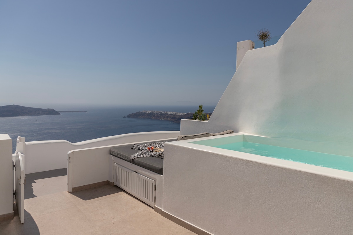An outdoor terrace features a heated plunge pool surrounded by a smooth, white wall. A spacious sunbed provides relaxation space, offering unobstructed views of the caldera and the horizon.