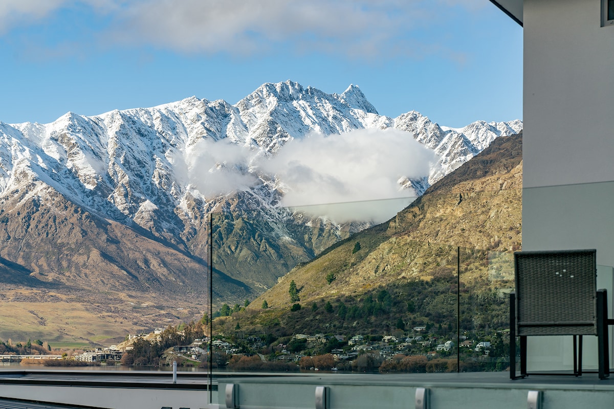 A balcony view showcases the breathtaking snow-capped Remarkables mountain range, partially covered by wispy clouds. A glass railing offers clear sightlines to the mountains and the valley below, enhancing the serene atmosphere of the lakeside retreat.