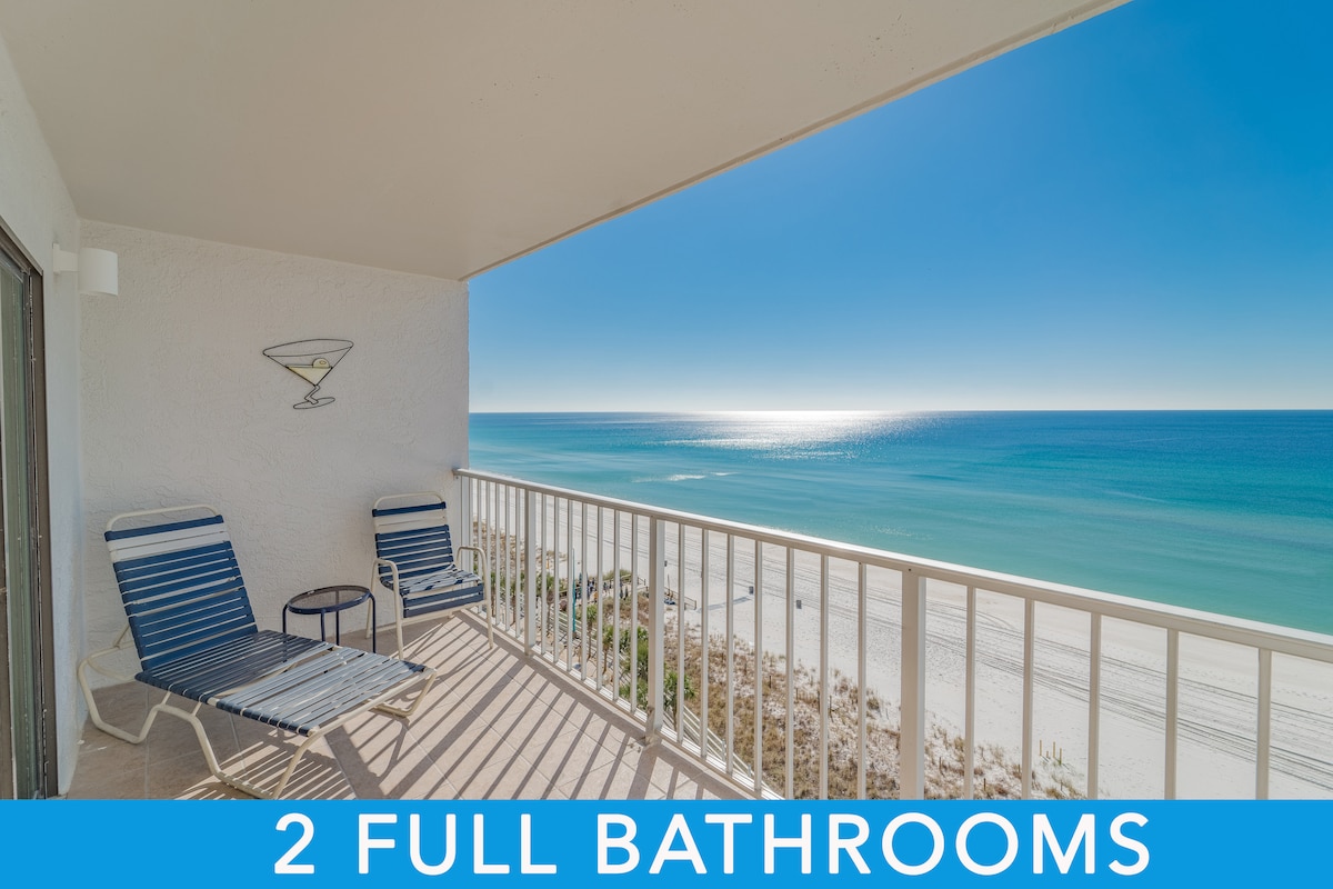 A private balcony is visible, featuring two lounge chairs and a small table, overlooking the serene beach and clear blue water. The expansive ocean view extends to the horizon, with sunlight glimmering on the surface. The space is designed for relaxation and enjoying scenic coastal vistas.