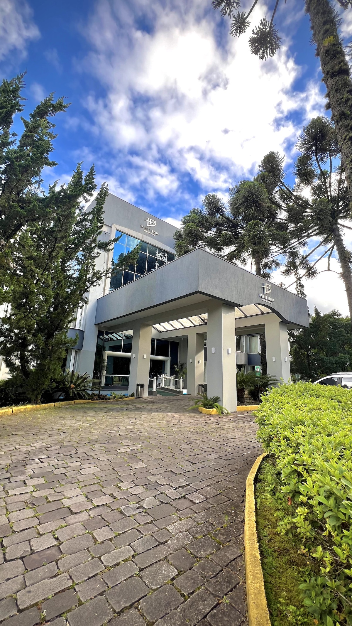 The modern building showcases large glass windows and a covered entrance. Lush greenery and neatly trimmed hedges frame the path leading to the door, while tall trees rise alongside the structure under a partly cloudy sky.