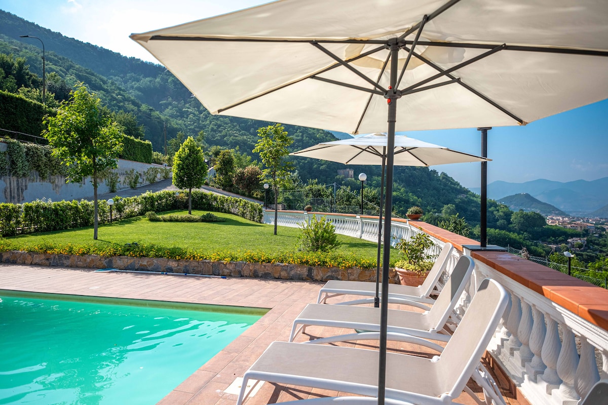 A sparkling swimming pool is surrounded by lounge chairs and large umbrellas, providing shaded seating. Lush green gardens and trees are visible in the background, with gentle hills complementing the serene landscape.