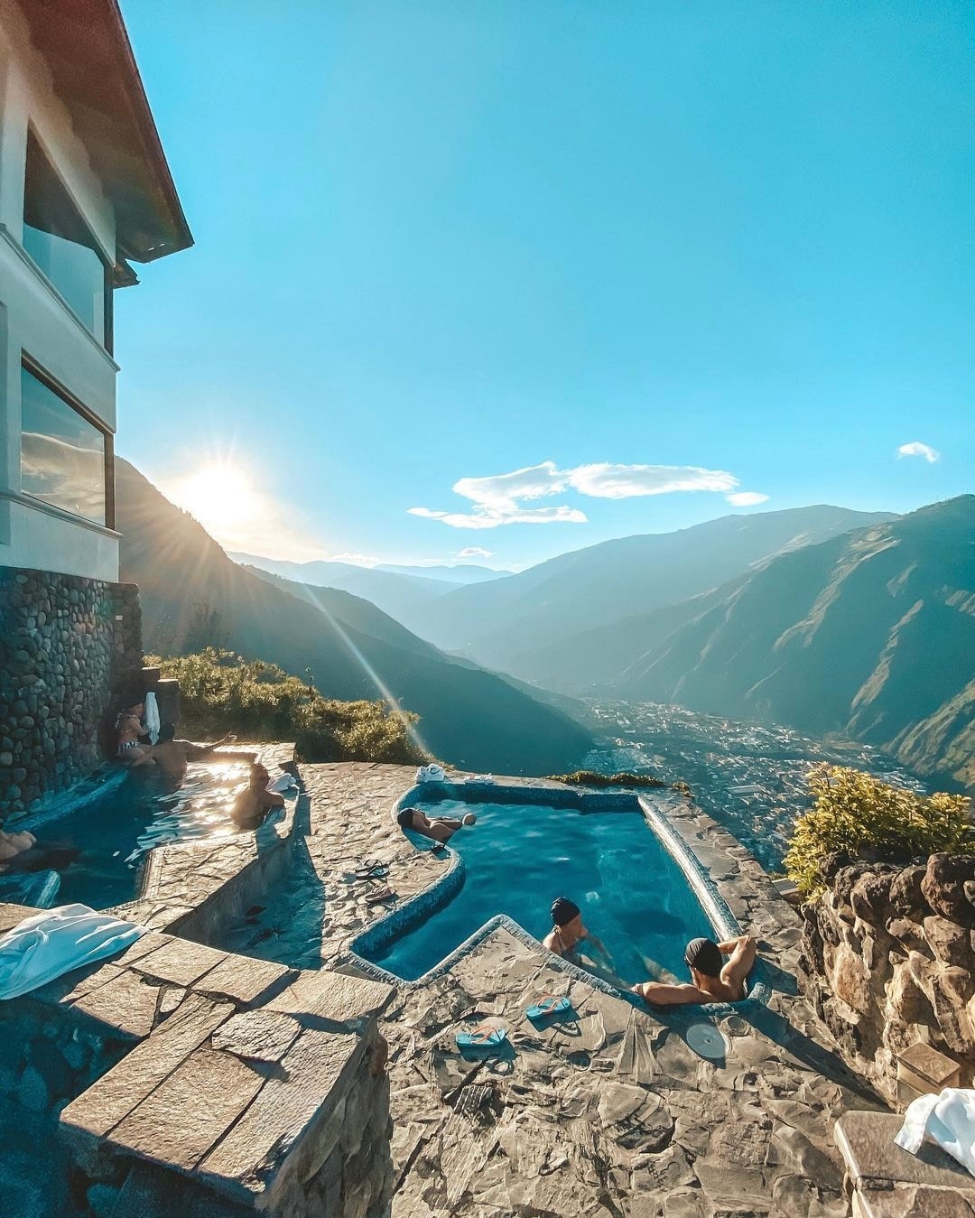Two outdoor hot tubs are positioned on a stone terrace, overlooking a valley framed by mountains. Sunlight bathes the scene, reflecting off the water. People are seen relaxing in the hot tubs, enhancing the tranquil atmosphere.