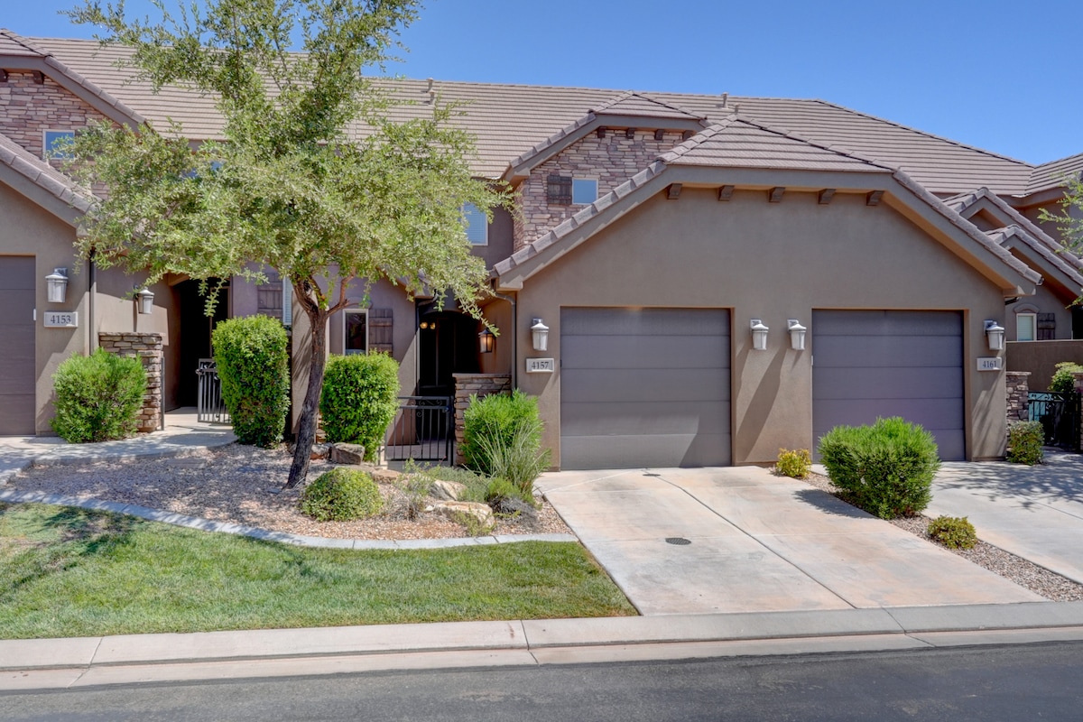 The exterior of a well-maintained multi-unit home is displayed, featuring stone accents and light brown stucco. Two garage doors are visible, flanked by green shrubs and small trees. A clear blue sky complements the warm tones of the structure.