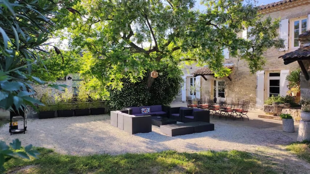 A spacious outdoor area is shaded by a large tree, featuring a comfortable seating arrangement with dark cushions. Nearby, a dining set is visible, ideal for gatherings. Flower beds and greenery enhance the serene environment, while the stone facade of the house provides a rustic backdrop.