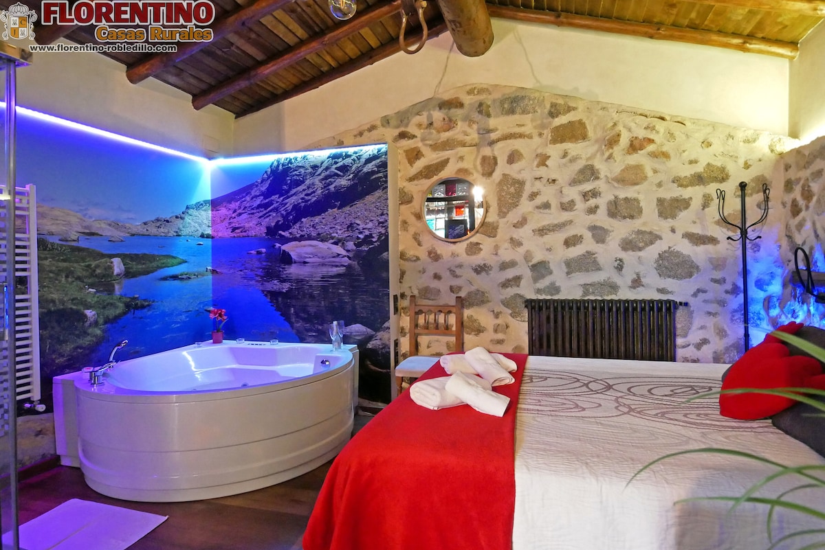 A modern sleeping area features a spacious bed adorned with red linens. A private jacuzzi is positioned nearby, complemented by a wall mural depicting a serene landscape. Exposed stone walls and wooden beams enhance the rustic charm of the room.