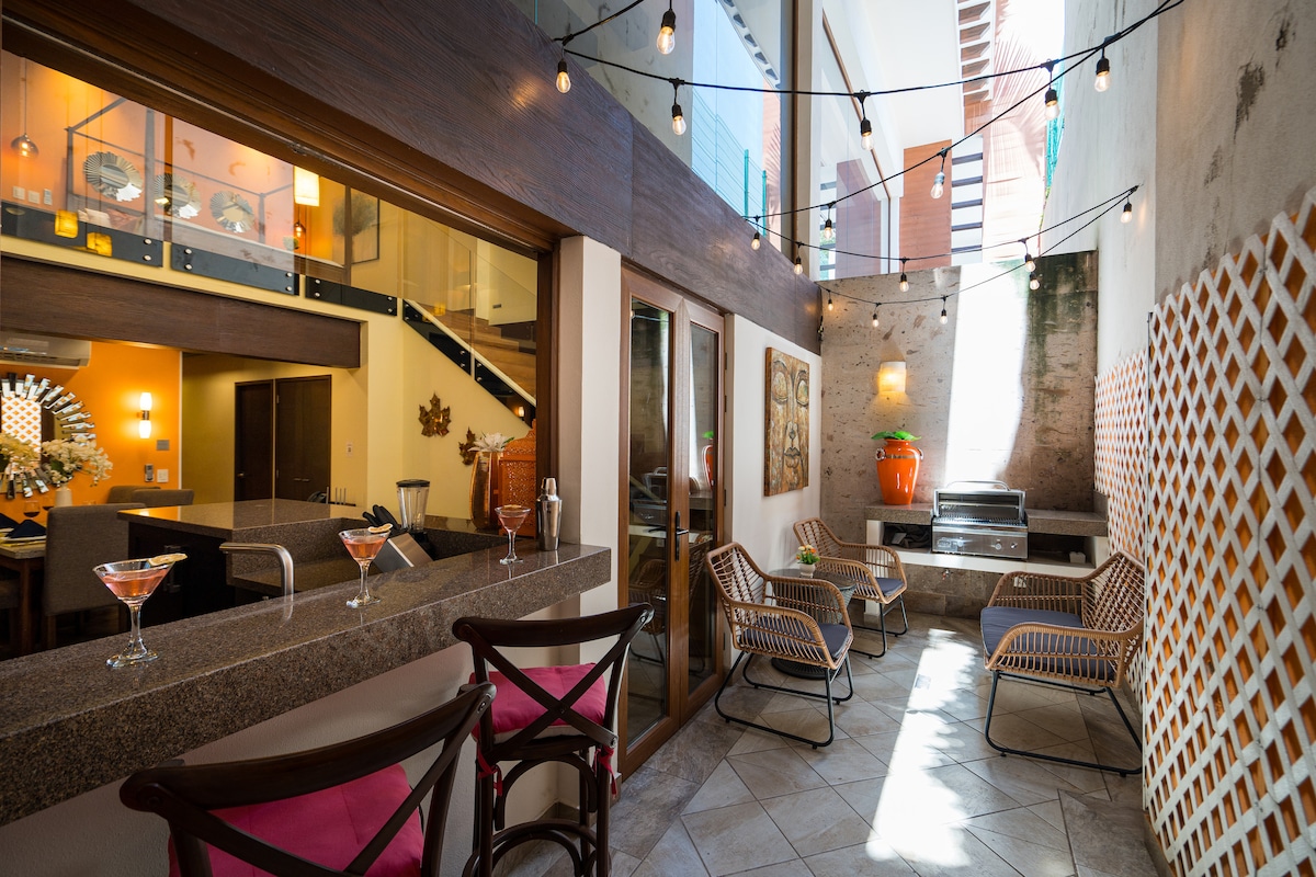 An inviting outdoor space is highlighted by warm string lights and modern furniture. Two chairs are positioned beside a stone wall, and a small bar area holds two drinks. A glimpse of the interior is visible through the large glass doors, showcasing a cozy and open layout.