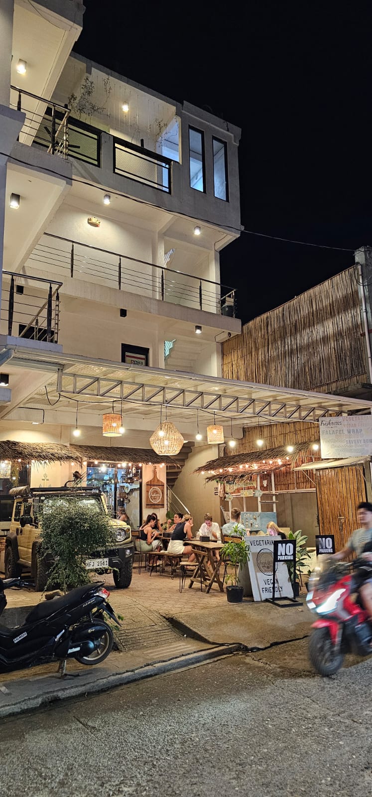 A multi-story building is illuminated at night, showcasing a bustling outdoor dining area with patrons seated at wooden tables. Hanging lights create a cozy ambiance, while a nearby street shows parked motorcycles and a flow of pedestrians. The overall scene captures the lively atmosphere of the local dining scene.