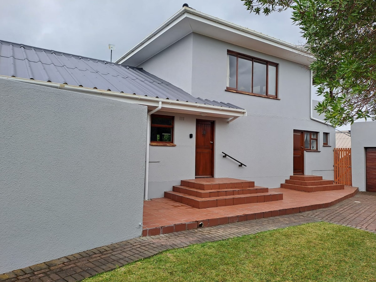 A two-story house features a grey exterior with a pitched roof. Steps lead to the front door, surrounded by a tiled patio. The entryway is flanked by wooden doors, and a neatly manicured lawn is visible in the foreground.