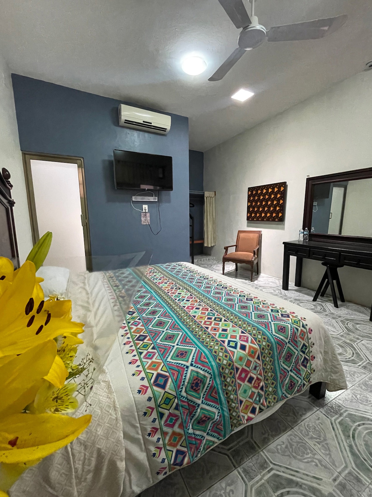 A spacious room features a King Size bed adorned with a colorful patterned blanket, enhancing visual appeal. A wooden chair and a functional desk are positioned nearby. Natural light fills the space from a window, while a wall-mounted TV and air conditioning unit are also present.