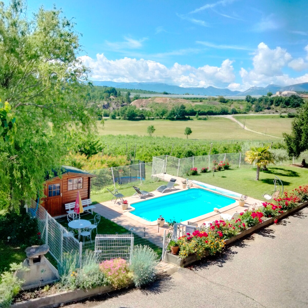 An outdoor pool is surrounded by a well-maintained garden featuring colorful flowers. A wooden shed is visible near the pool, and lounging chairs are positioned around the area. The expansive landscape showcases greenery and distant hills under a clear blue sky.