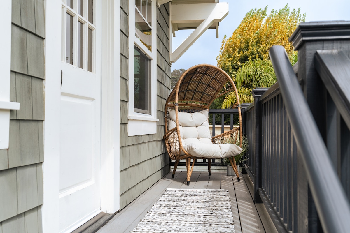 A cozy outdoor seating area is presented on the deck, featuring a rattan chair with soft cushions. A small, decorative rug lies in front, while greenery is visible in the background, creating a peaceful setting near the entrance.
