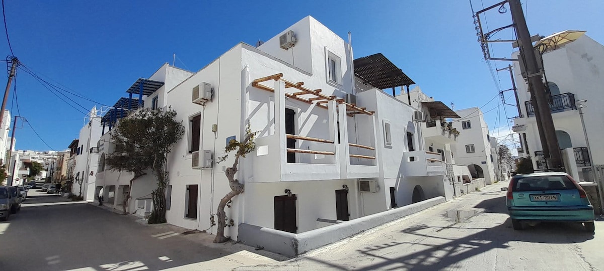 A whitewashed building with multiple levels is observed, featuring balconies and air conditioning units. The street is lined with greenery and provides a clear view of the architecture, soaking in natural light under a blue sky.