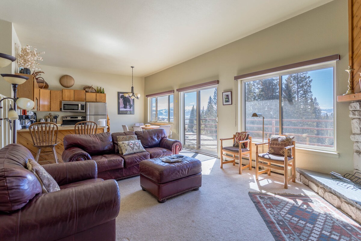 A spacious living area features a comfortable leather couch and rustic wooden chairs, with natural light flooding in from large windows. A view of the mountains is visible beyond the balcony. The adjacent kitchen area and dining space can be seen in the background.