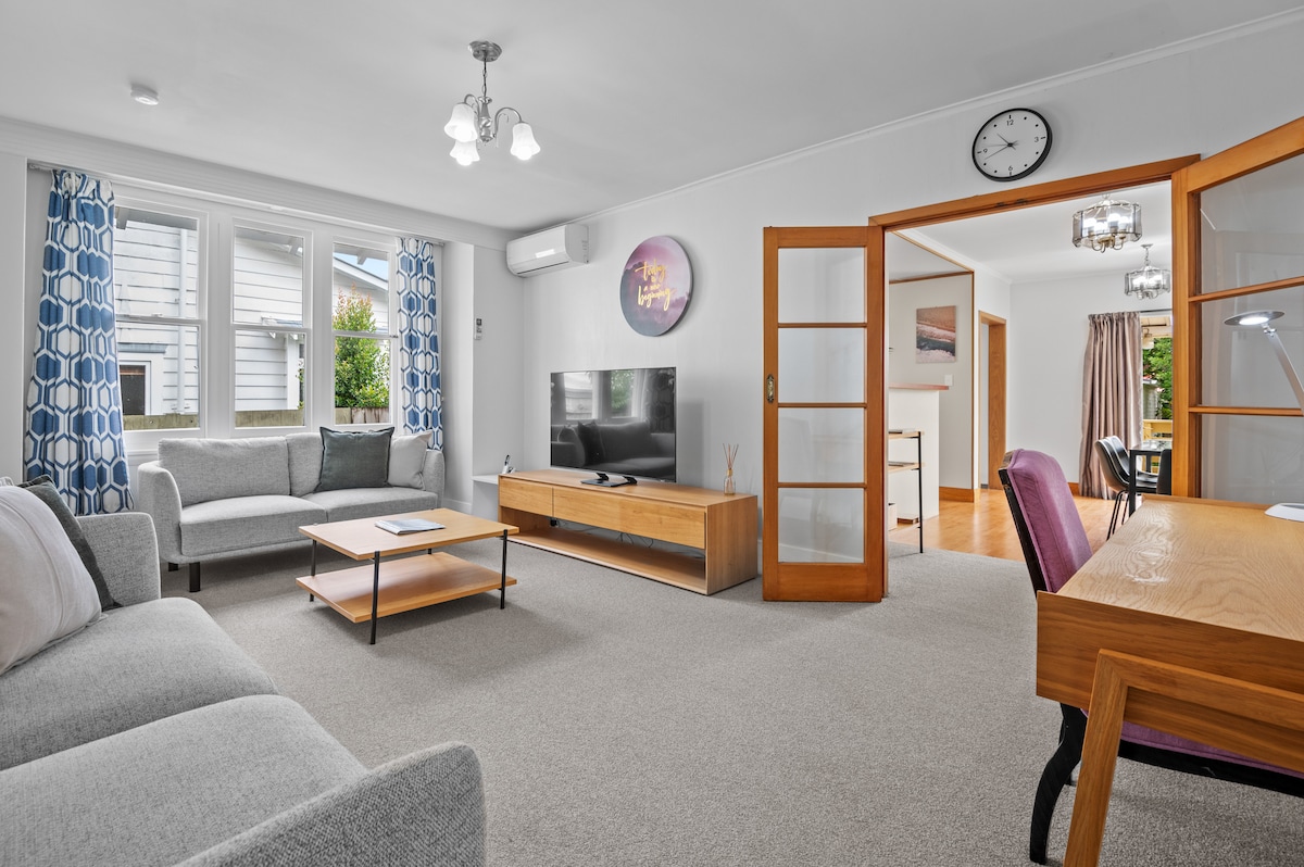 A spacious living area features two light gray sofas and a wooden coffee table positioned centrally. A wall-mounted television is visible, alongside a wooden desk with a purple chair. Natural light floods in through large windows with patterned curtains, enhancing the welcoming environment.