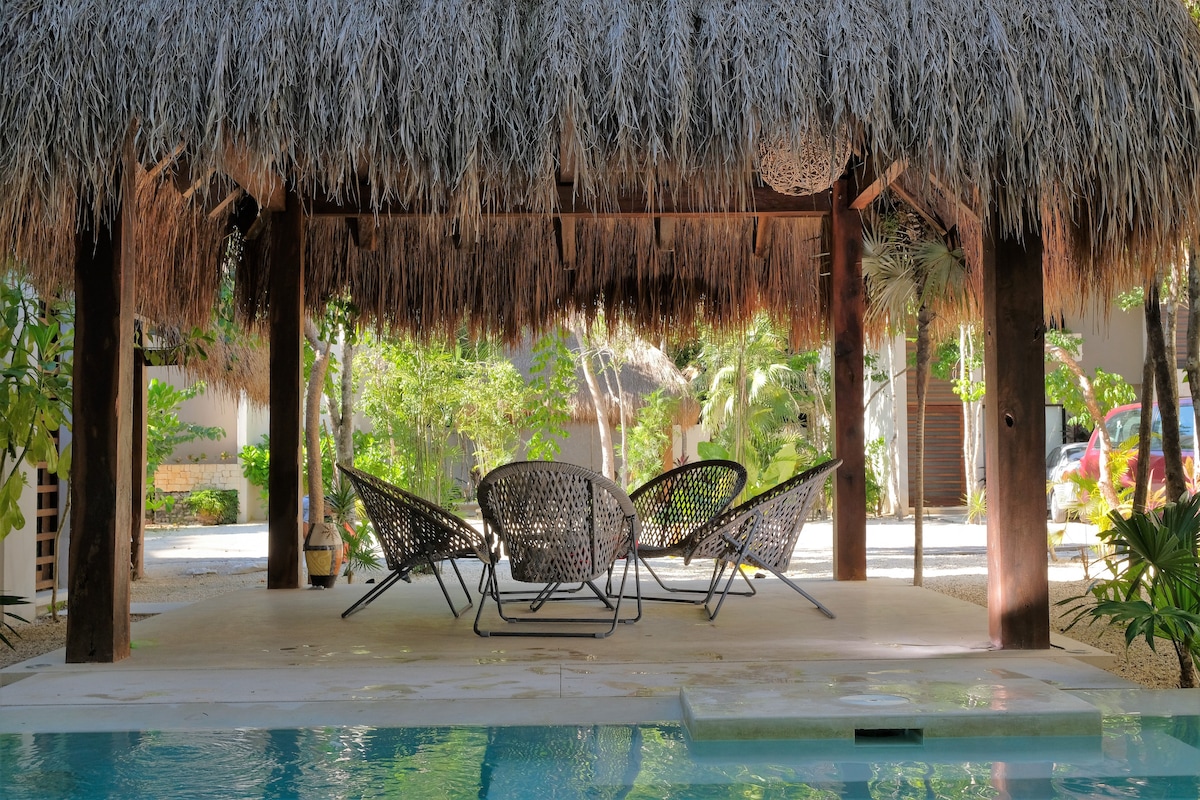 A shaded outdoor area features four comfortable woven chairs arranged under a thatched roof, surrounded by lush greenery. A tranquil pool reflects the natural surroundings, enhancing the serene ambiance of this inviting space.