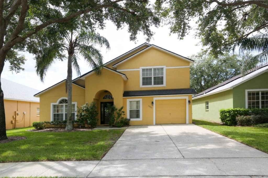 A two-story villa is presented with a bright yellow exterior, surrounded by lush greenery and palm trees. A driveway leads to the entrance, featuring a decorative archway and a single-car garage. Neighboring properties are visible, with one displaying a complementary green hue.