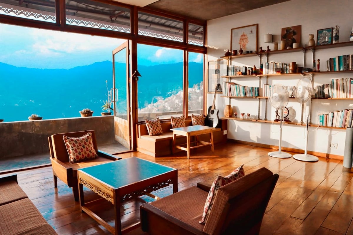 A spacious living area features large windows offering views of the mountains. Wooden furniture, including low tables and comfortable seating, is arranged around the room. Bookshelves line one wall, showcasing various books and decorative items. Natural light brightens the cozy, wooden interior.