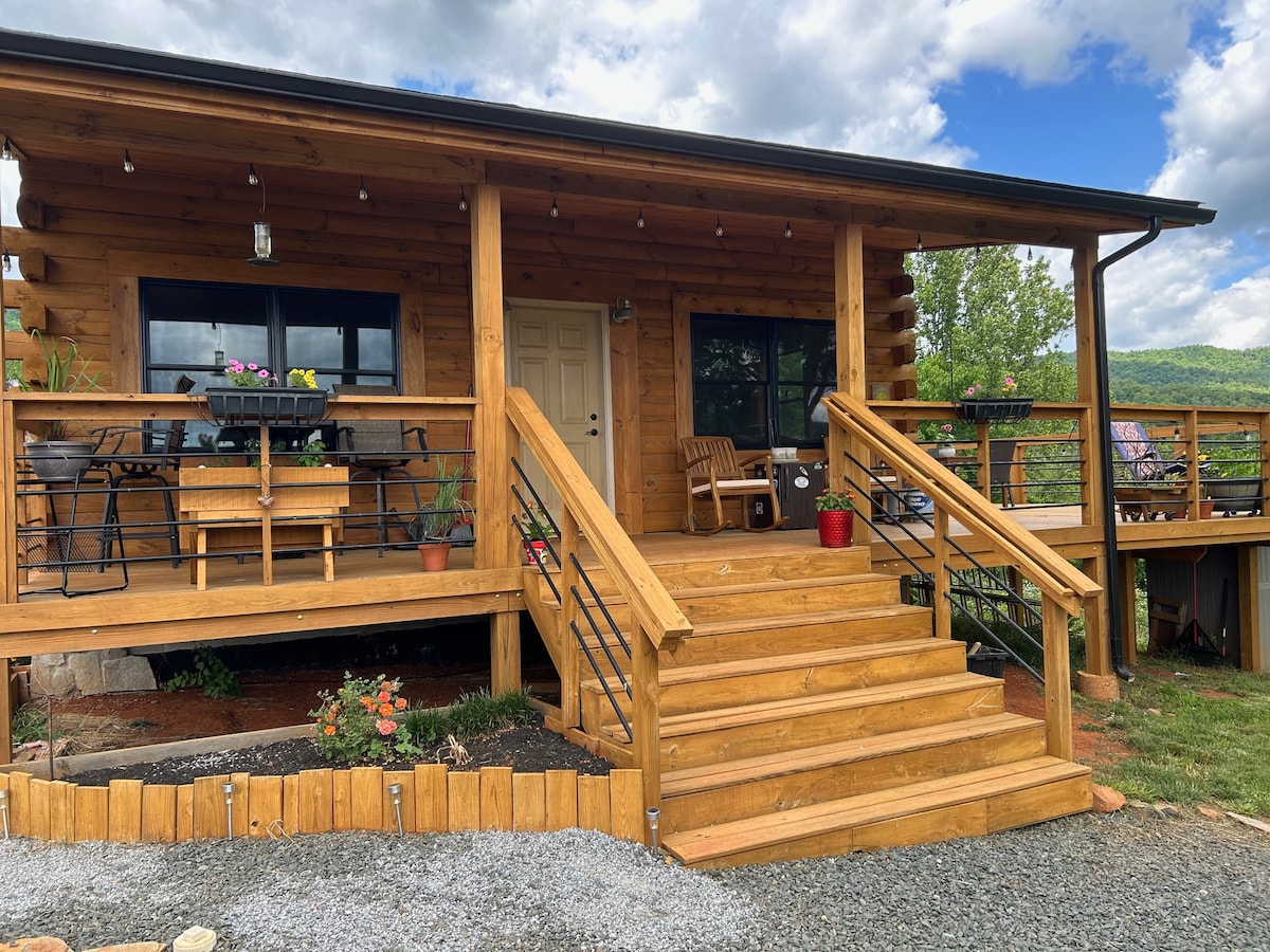 The exterior of the log cabin showcases a welcoming entrance with wooden stairs leading to a front door. A wraparound deck features seating areas and potted plants, with views of the surrounding landscape and a cloudy sky.