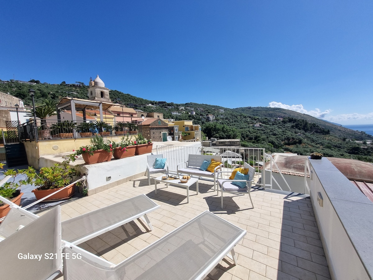 A spacious outdoor terrace features multiple lounge chairs and a dining area. Potted plants adorn the railing, offering greenery against the backdrop of the scenic hillside and coastline. The clear sky enhances the tranquil setting, making it ideal for outdoor relaxation.
