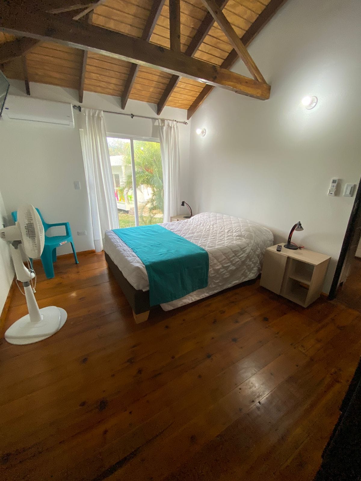 A bright sleeping area is enhanced by wooden beams overhead and natural light filtering through a large sliding door. A spacious bed is made with white linens and a turquoise throw. A nightstand with a lamp and a fan is positioned next to the bed, and a chair occupies a corner.
