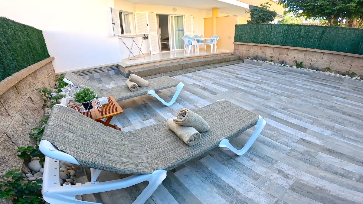 A private terrace features two loungers with neatly arranged towels. A small wooden table is positioned beside the loungers. A partial view of an opening leads to a light-filled indoor area, surrounded by greenery and decorative stones, creating a relaxing outdoor space.