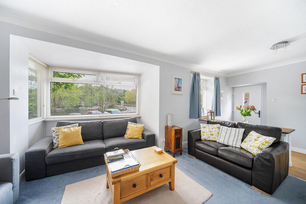 A spacious living room features two comfortable sofas, one in dark leather and the other in fabric, both adorned with decorative cushions. A wooden coffee table occupies the center, surrounded by natural light from large windows with curtains. A dining area is visible in the background.