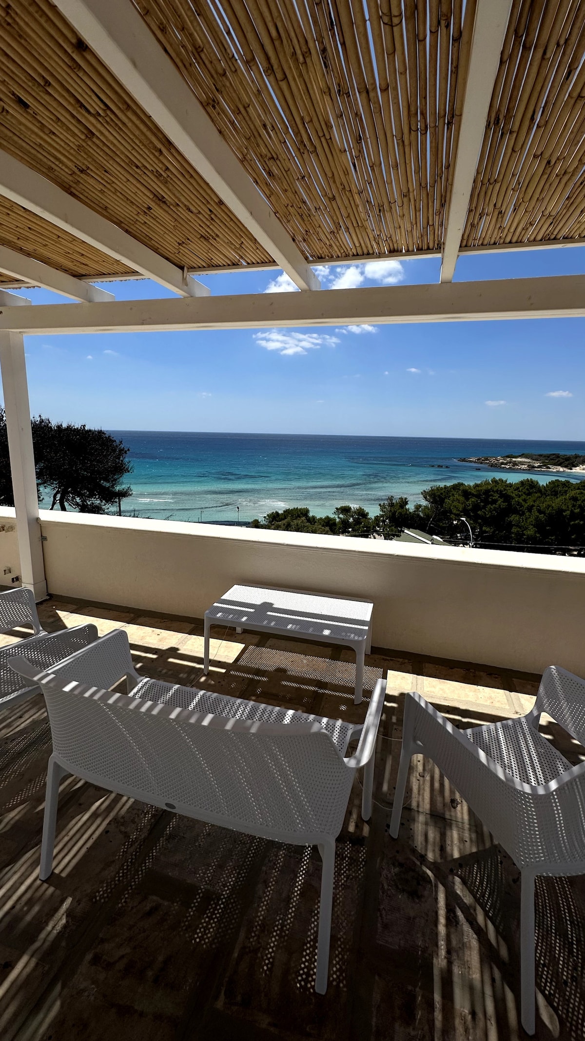 A terrace with a thatched roof provides a view of the blue sea and horizon. White outdoor chairs surround a small table, inviting relaxation. The natural scenery includes sandy beaches and greenery, highlighting the peaceful coastal ambiance.