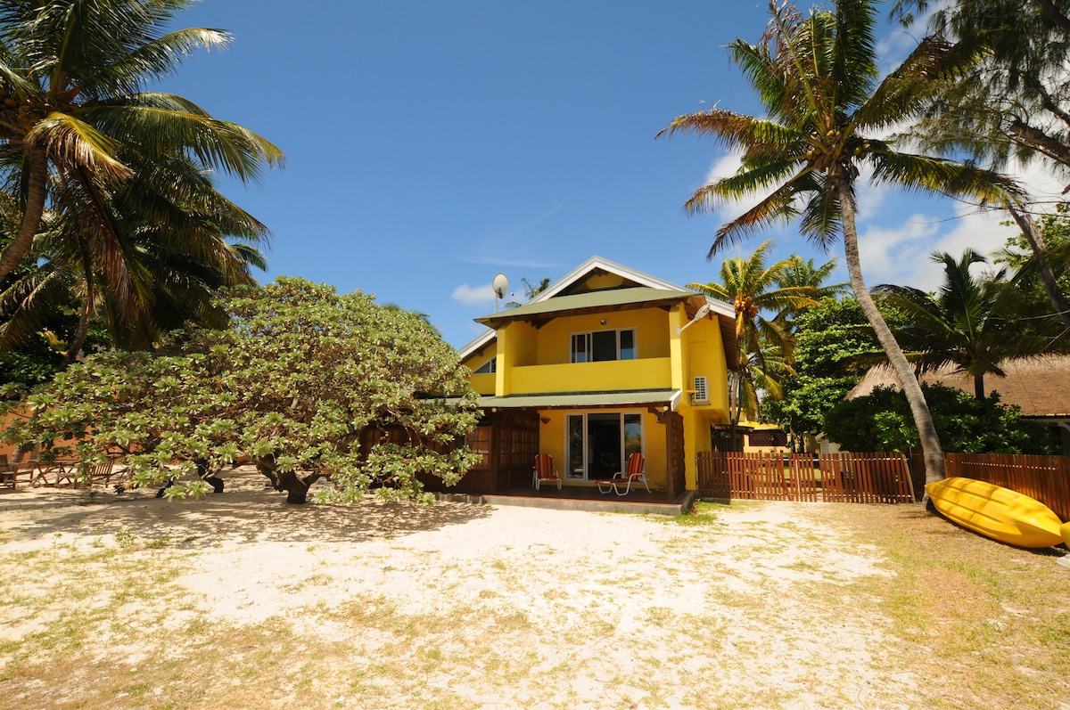 A two-story villa is surrounded by lush greenery and palm trees, set against a clear blue sky. The bright yellow exterior stands out, and the terrace features chairs. A sandy area leads toward the beach, creating a tranquil outdoor space.