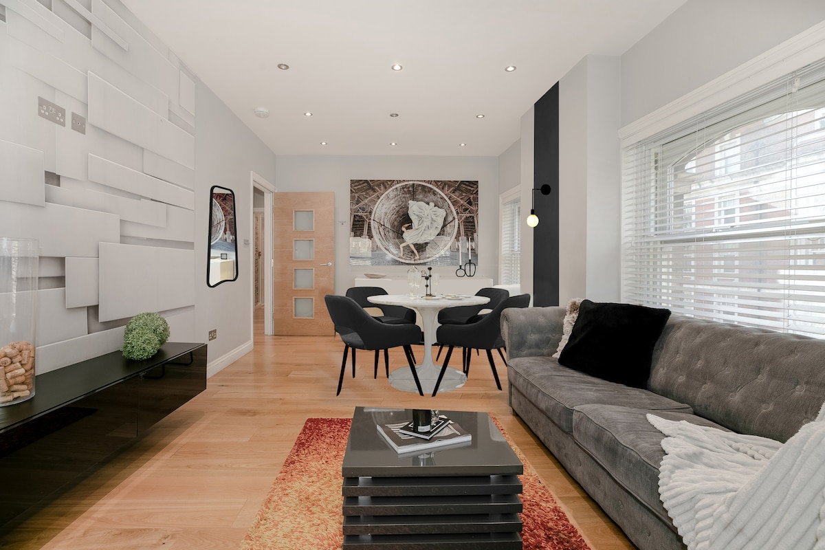 A modern living space features sleek furnishings with a plush grey sofa and two stylish black chairs. A glass coffee table sits atop a warm red and orange area rug. A large piece of art decorates the wall, complemented by natural light from the window dressed with subtle blinds.