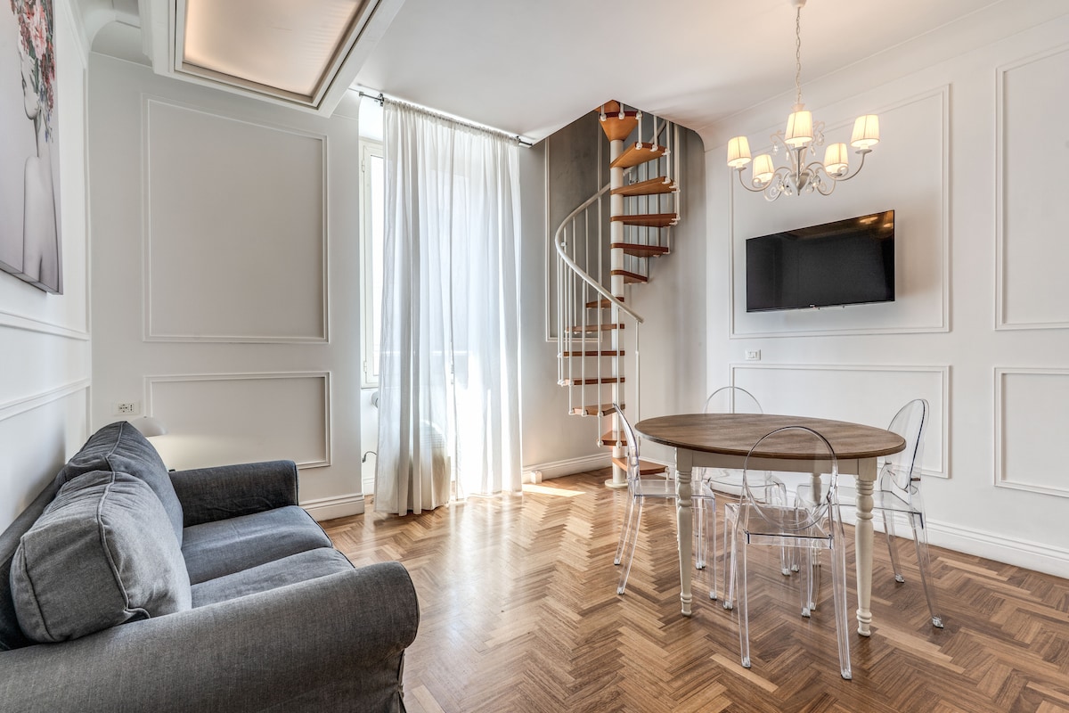 A living area is showcased with herringbone wooden flooring and large windows that allow natural light to fill the space. A cozy sofa is positioned near a round dining table surrounded by transparent chairs. A spiral staircase leads to the upper level, complemented by a modern light fixture.