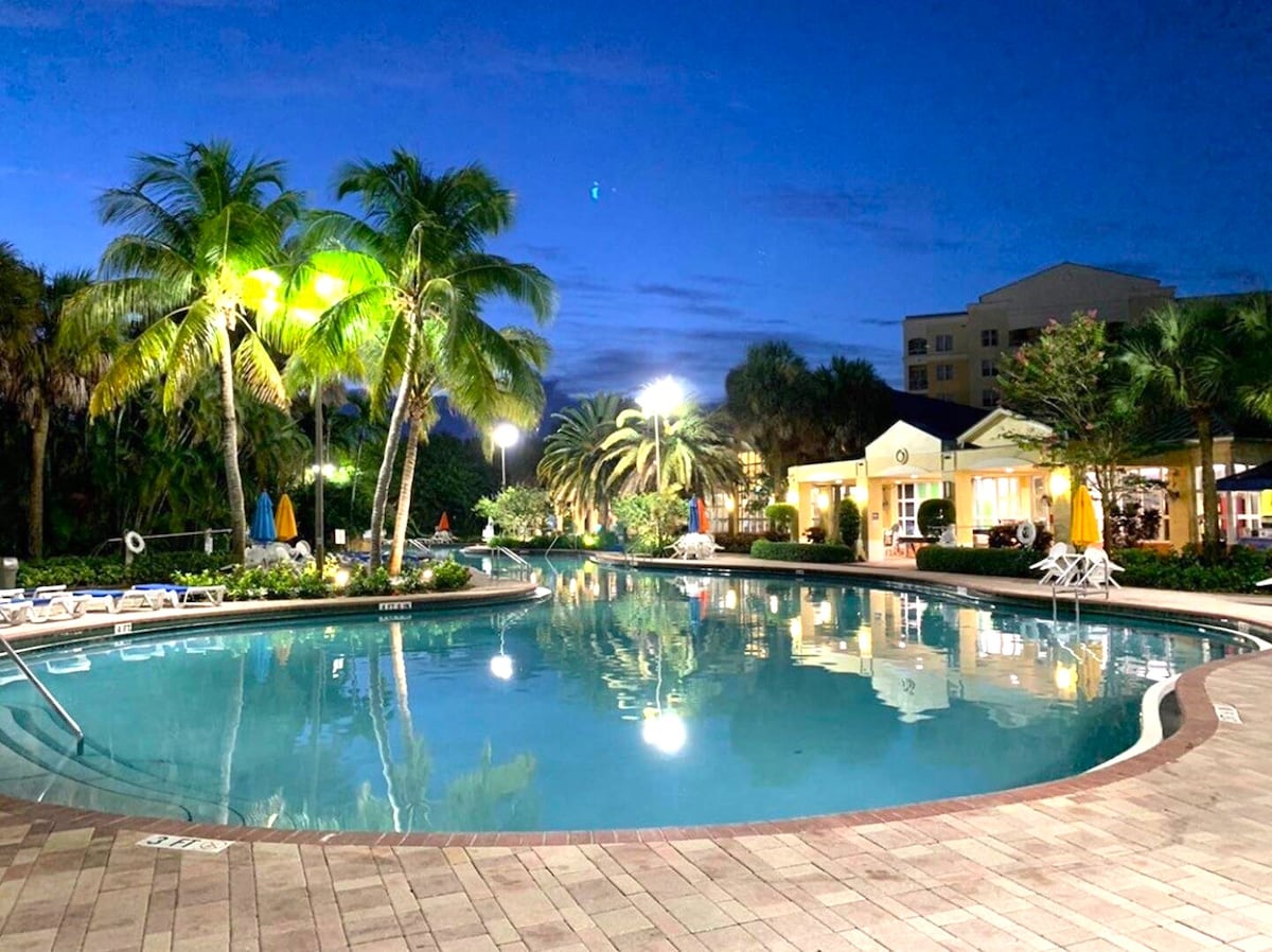 The pool area is illuminated by warm lights, surrounded by tropical palm trees. Lounge chairs line the pool's edge, with colorful umbrellas providing shade. The serene water reflects the evening sky, creating a tranquil atmosphere, while a clubhouse is visible in the background.