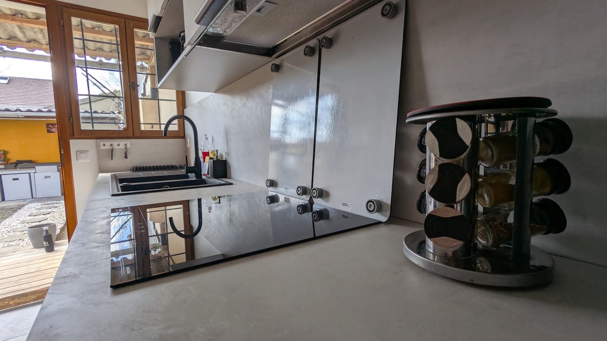 A modern kitchen is displayed with a sleek countertop and minimalistic design. A black glass cooktop is positioned beside a sink, with organizational items visible on the counter. A circular spice rack holds several jars, adding practical storage to the space.