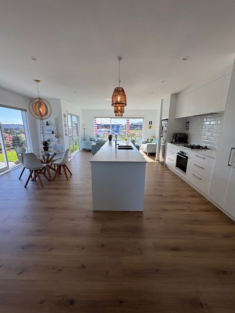 An open living space features a modern kitchen with a central island and barstools. A dining area with a round table and white chairs is positioned near a window, allowing natural light to flow in, while glass doors lead to a garden view.