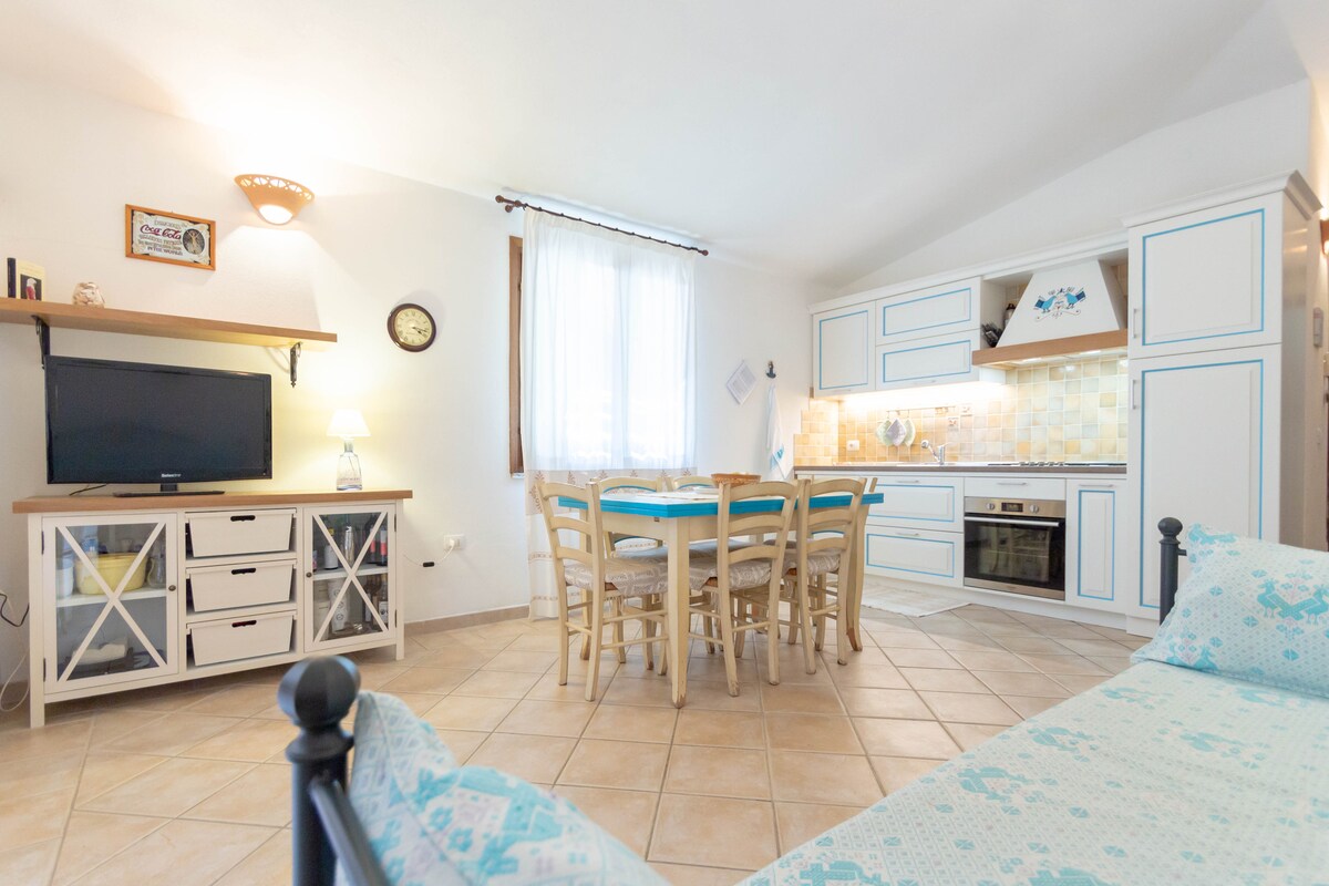 A bright living area is highlighted by a spacious dining table surrounded by six wooden chairs. A well-equipped kitchen, featuring light cabinetry and modern appliances, is visible in the background. Soft natural light enters through a window, illuminating the neutral tiled floor.