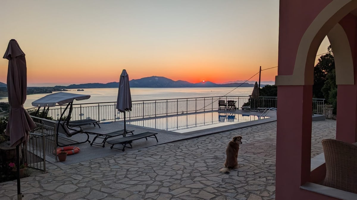 A tranquil sunset view overlooks the serene water, featuring a swimming pool and lounge chairs on a paved terrace. Umbrellas are positioned near the pool area, while a dog is seated on the stone path, adding a sense of calm to the scene.