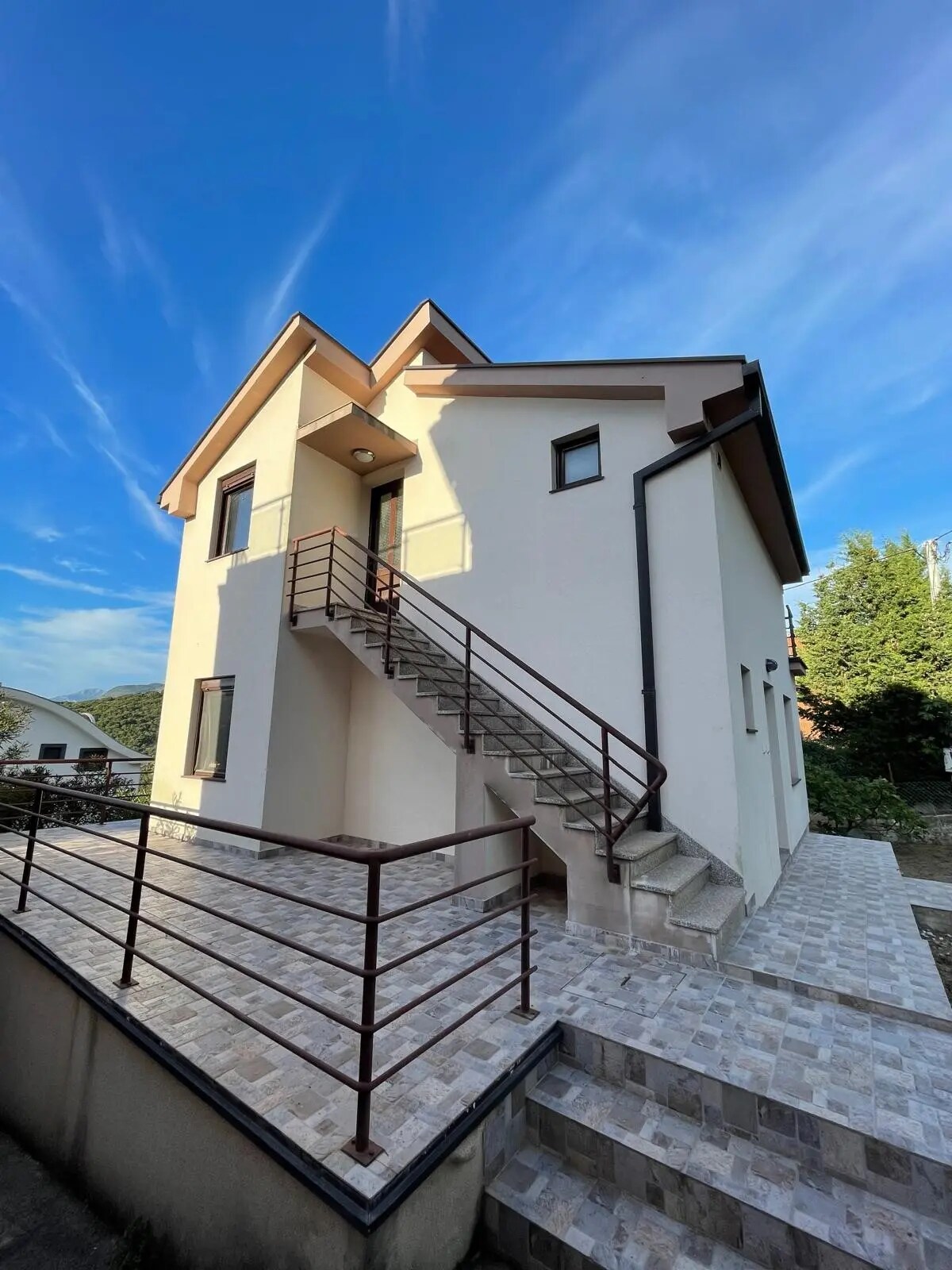 A two-story house is depicted, featuring a modern exterior with a blend of light-colored walls and a sloped roof. A set of stairs leads to the entrance, and a railing runs alongside the path, providing safety. The background showcases greenery under a clear blue sky.