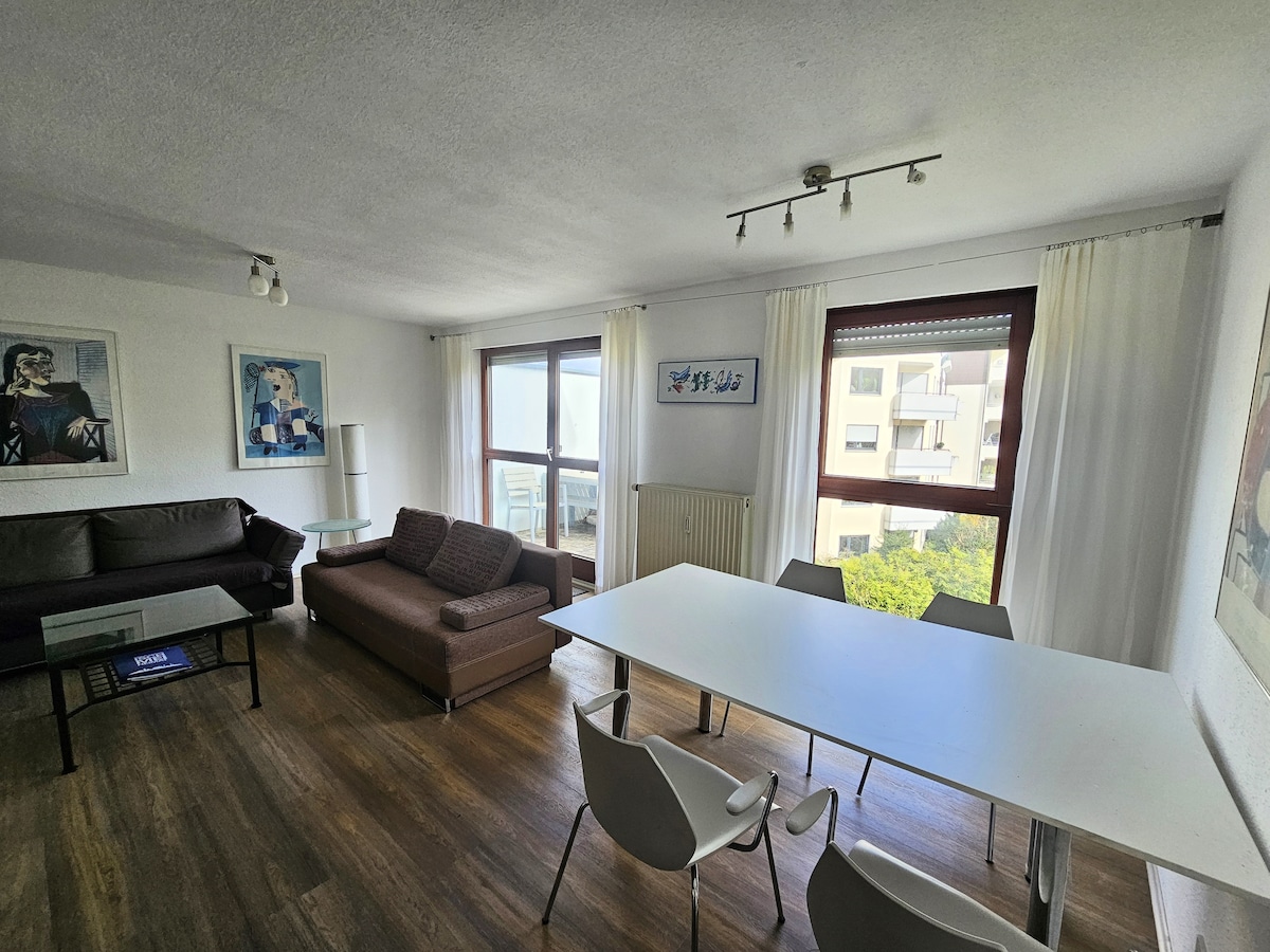 A bright living area is highlighted by large windows offering natural light and views. A comfortable sofa is positioned next to a sleek armchair. A modern dining table with four chairs sits centrally, surrounded by minimalistic artwork on the walls.