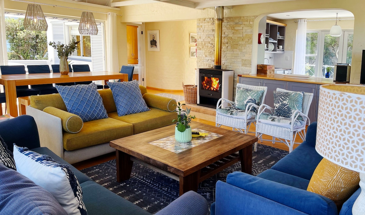 A spacious living area features a combination of comfortable seating arrangements, including a mix of colorful sofas and armchairs. A wooden coffee table sits in the center, adorned with a small potted plant. A cozy wood-burning fireplace adds warmth to the inviting atmosphere.