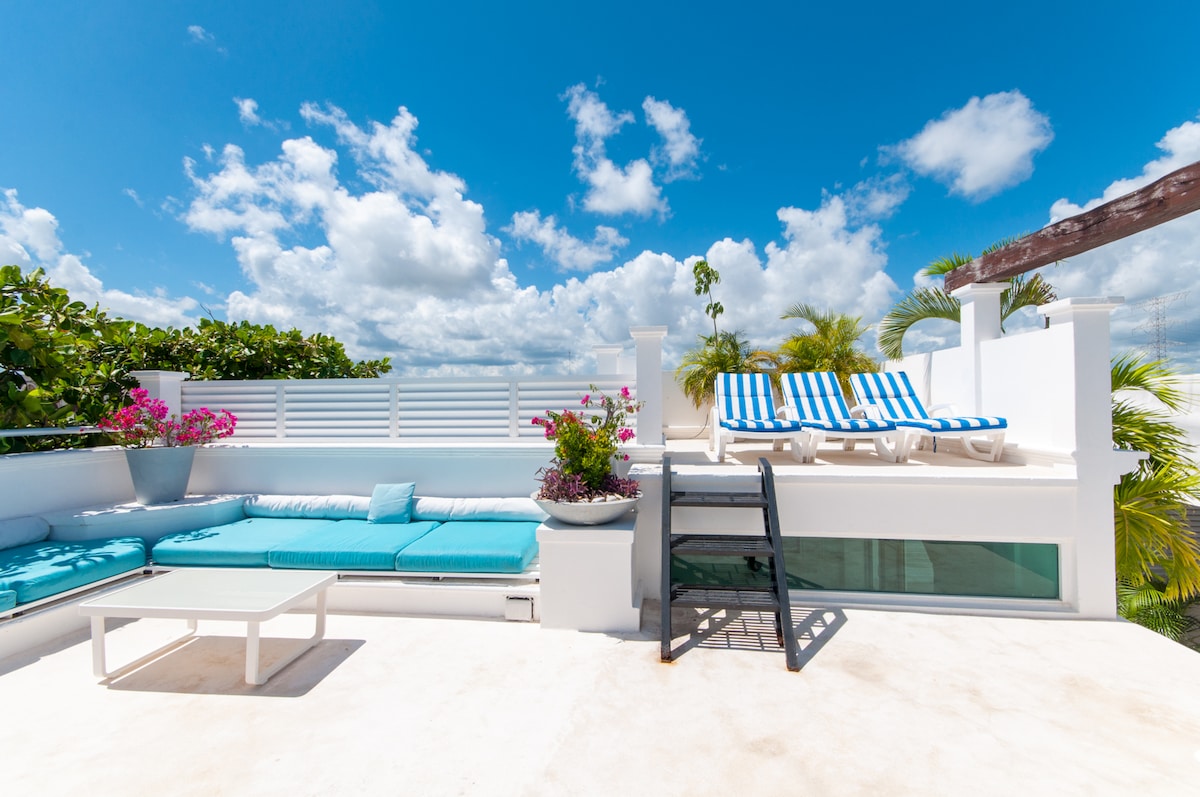 The rooftop terrace features stylish lounge chairs and a spacious daybed adorned with turquoise cushions. Vibrant plants in pots add a splash of color, while a clear blue sky with fluffy clouds creates a serene backdrop for relaxation.
