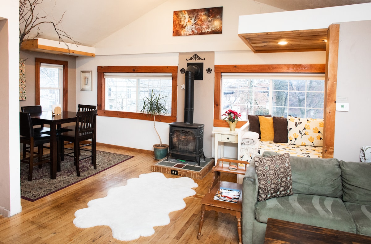 The open living space features vaulted ceilings with large windows allowing natural light, a wood stove, and a cozy day bed. A dining area with a four-person table is adjacent to a comfortable seating area, creating an inviting atmosphere for relaxation.