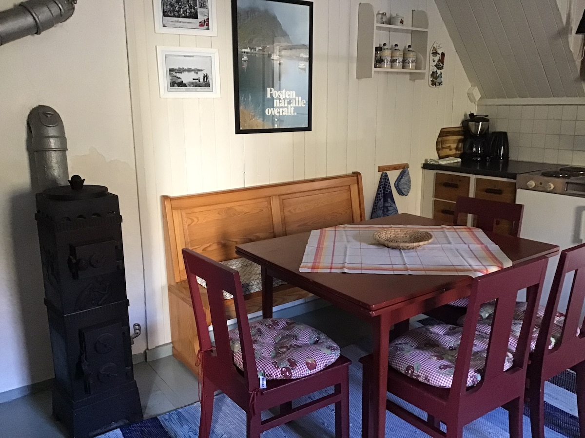 A dining area features a wooden table surrounded by four red chairs with cushioned seats. An antique wood stove stands nearby, complemented by a small shelf holding pantry items. The walls are adorned with framed photographs, creating a cozy setting.